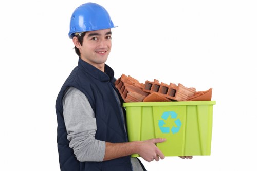 A young male waste management worker wearing a blue safety helmet and a dark blue vest over a grey long-sleeved shirt is holding a bright green recycling bin with a blue recycling symbol on the front. The bin is filled with red clay bricks that are stacked and protrude slightly from the top. Behind him is a plain white background, highlighting the focus on the individual and the bin. The worker is smiling subtly and appears to be preparing for rubbish removal services in Kensington, suited for dealing with construction debris or general waste disposal relevant to local commercial waste management needs. The image emphasizes the careful handling of building materials and the importance of proper waste segregation in an urban environment, supporting services offered by Commercial Waste Kensington in the town or postcode area around Kensington.